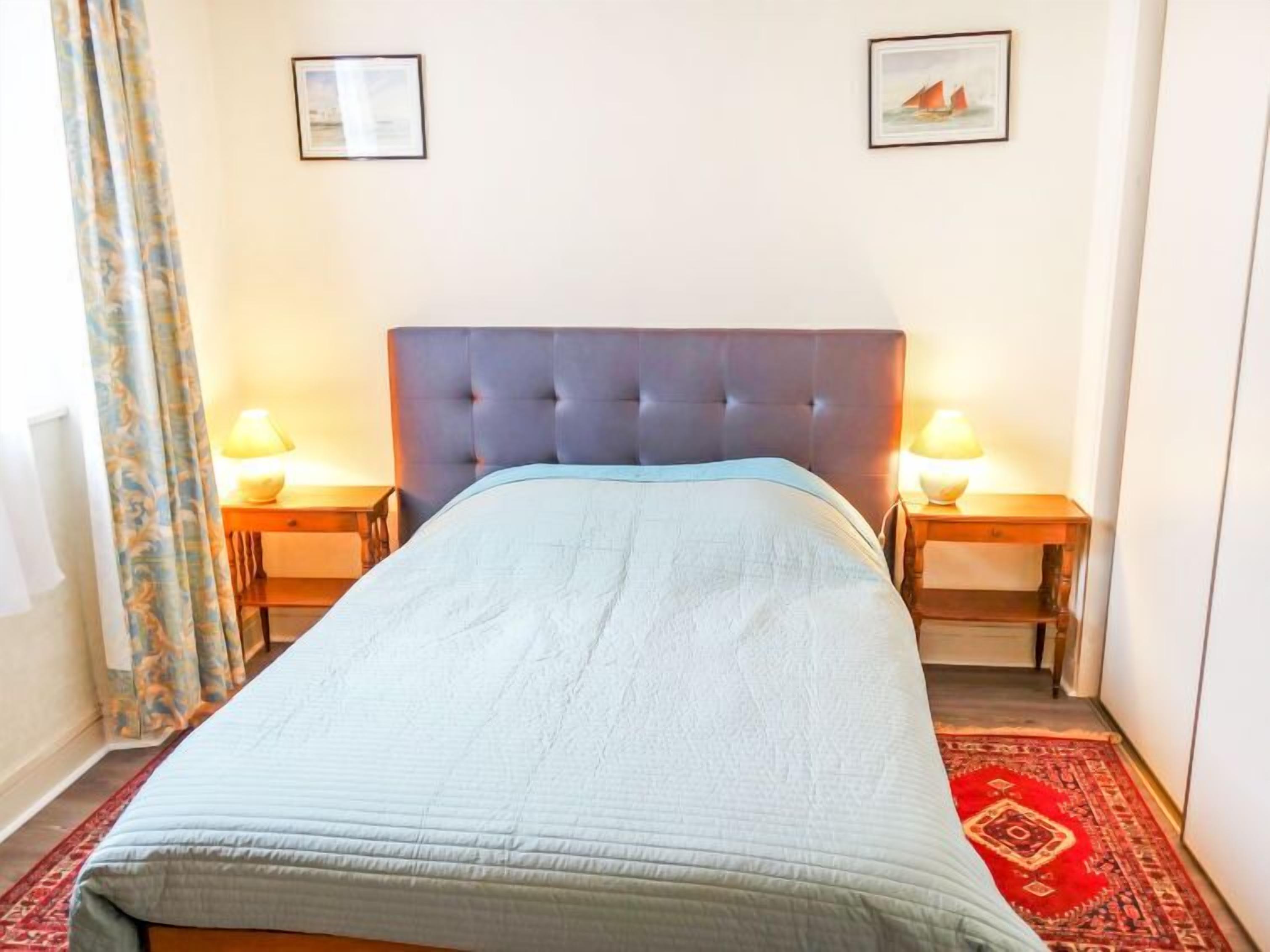 A neatly made bed with a blue headboard, two bedside lamps, and a red patterned rug.