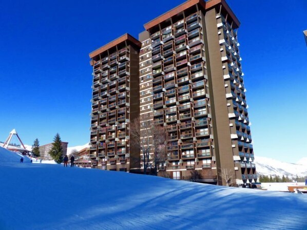 Un edificio de gran altura con múltiples balcones, un telesilla al fondo y un cielo azul despejado.