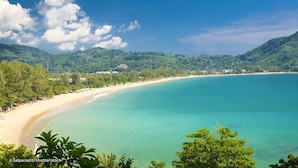 A coastal landscape with a sandy beach, clear turquoise waters, and lush green hills in the background. - Grand Kamala Falls (Kamala)