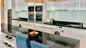 A modern kitchen with white cabinets, stainless steel appliances, a glass dining table, and a blue chair. - Grand Kamala Falls (Kamala)