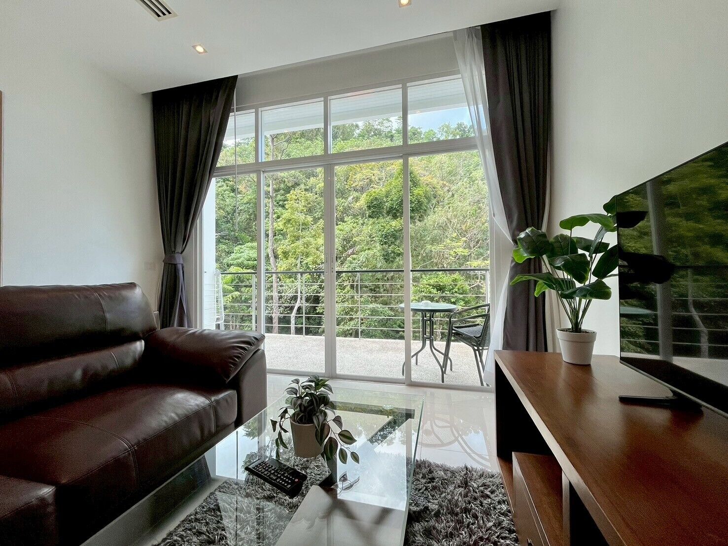 A modern living room with a leather sofa, a glass coffee table, a TV on a wooden stand, and a balcony with a table and chairs.