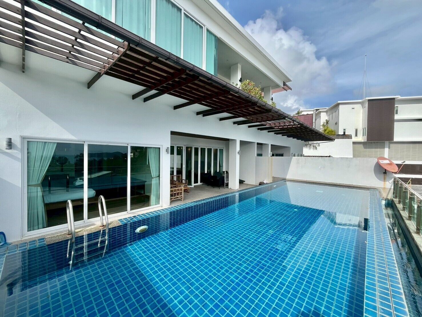A modern swimming pool with a clear blue tile design, adjacent to a white building with large windows and a covered patio area.