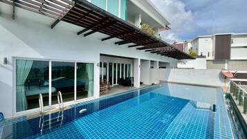 A modern swimming pool with a clear blue tile design, adjacent to a white building with large windows and a covered patio area.