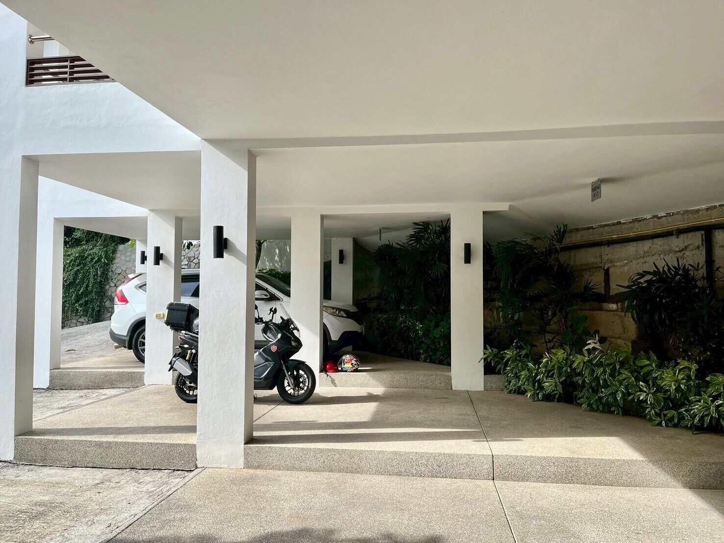 Un espace de stationnement couvert où sont garés une voiture blanche et un motocycle. Des marches en béton mènent à l’entrée, et l’ensemble est entouré de verdure.