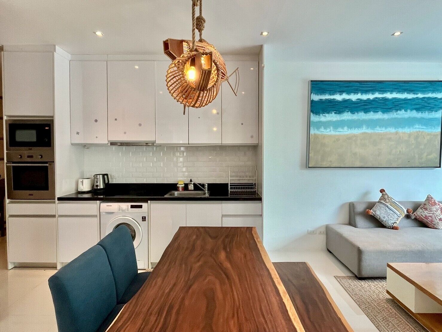 Eine moderne Küche mit weißen Schränken, einem Holztisch und einem großen Gemälde mit Strandmotiv.