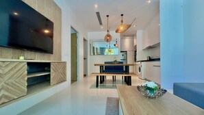 A modern living room with a flat-screen TV, wooden furniture, a dining area with a blue sofa, and a kitchen in the background. - Grand Kamala Falls (Kamala)