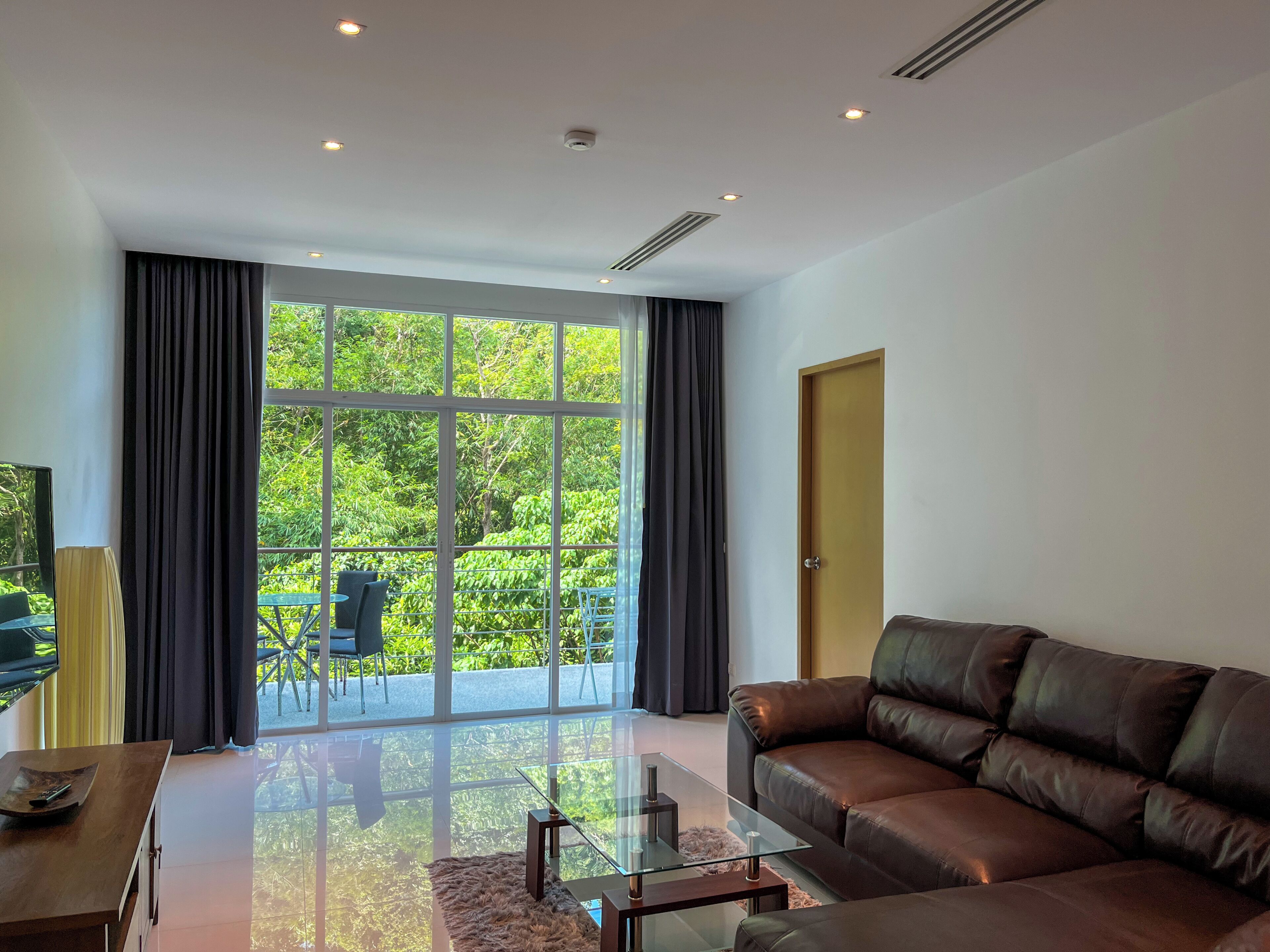 Una sala de estar moderna con un sofá de cuero, una mesa de centro de vidrio y un televisor sobre un mueble de madera. Amplias ventanas ofrecen vista a la vegetación exterior.