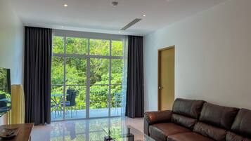 Una sala de estar moderna con un sofá de cuero, una mesa de centro de vidrio y un televisor sobre un mueble de madera. Amplias ventanas ofrecen vista a la vegetación exterior.