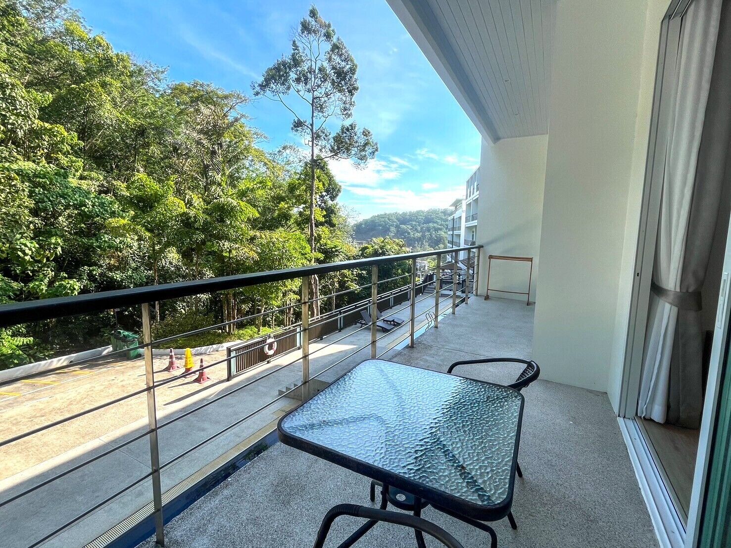 Un balcon avec une table et des chaises en verre, donnant sur une route et sur un espace verdoyant.