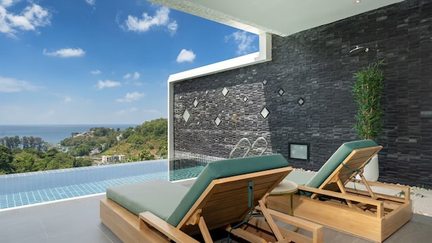 A modern outdoor lounge area with a pool, wooden loungers, a black stone wall, and a view of the sea. - Grand Kamala Falls (Kamala)