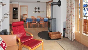 A cozy living room with a red leather chair, a dining area with a table and chairs, a wood-burning stove, and a kitchen in the background. - Panorama Apt. P by Interhome (Wengen)