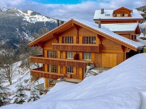 A wooden chalet with multiple balconies and a snow-covered roof, set against a mountainous backdrop.