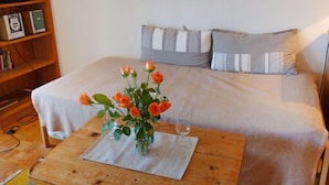 Une chambre à coucher avec un lit, des oreillers, une table de chevet en bois, un vase avec des fleurs et un téléviseur posé sur une étagère.