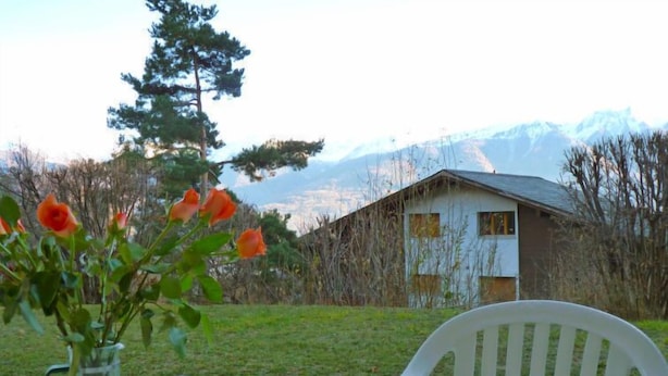 Une table avec un vase de fleurs et deux tasses, une chaise blanche, et une vue sur une maison et des montagnes en arrière-plan.