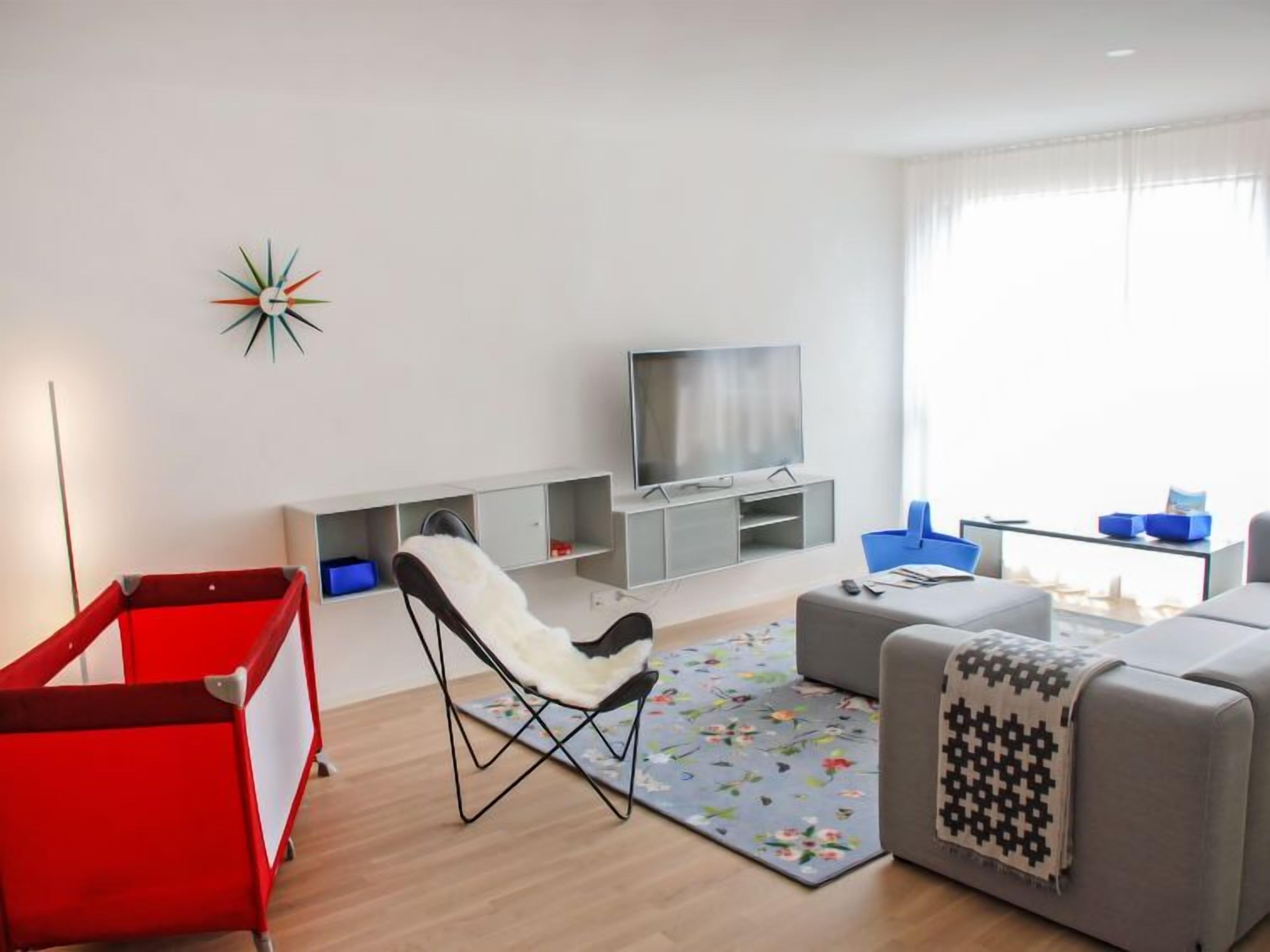 Uma sala de estar moderna com uma televisão, um cercado infantil vermelho, uma cadeira branca e um sofá cinza.