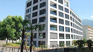Un edificio moderno de varios pisos con una fachada de ventanas y balcones, rodeado de vegetación y un cielo despejado.