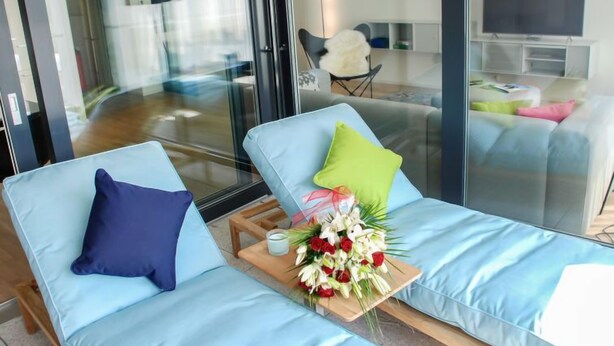 Una sala de estar moderna con dos sofás azul claro, una mesa de centro de madera con un arreglo floral, y un televisor de pantalla plana grande montado en la pared.