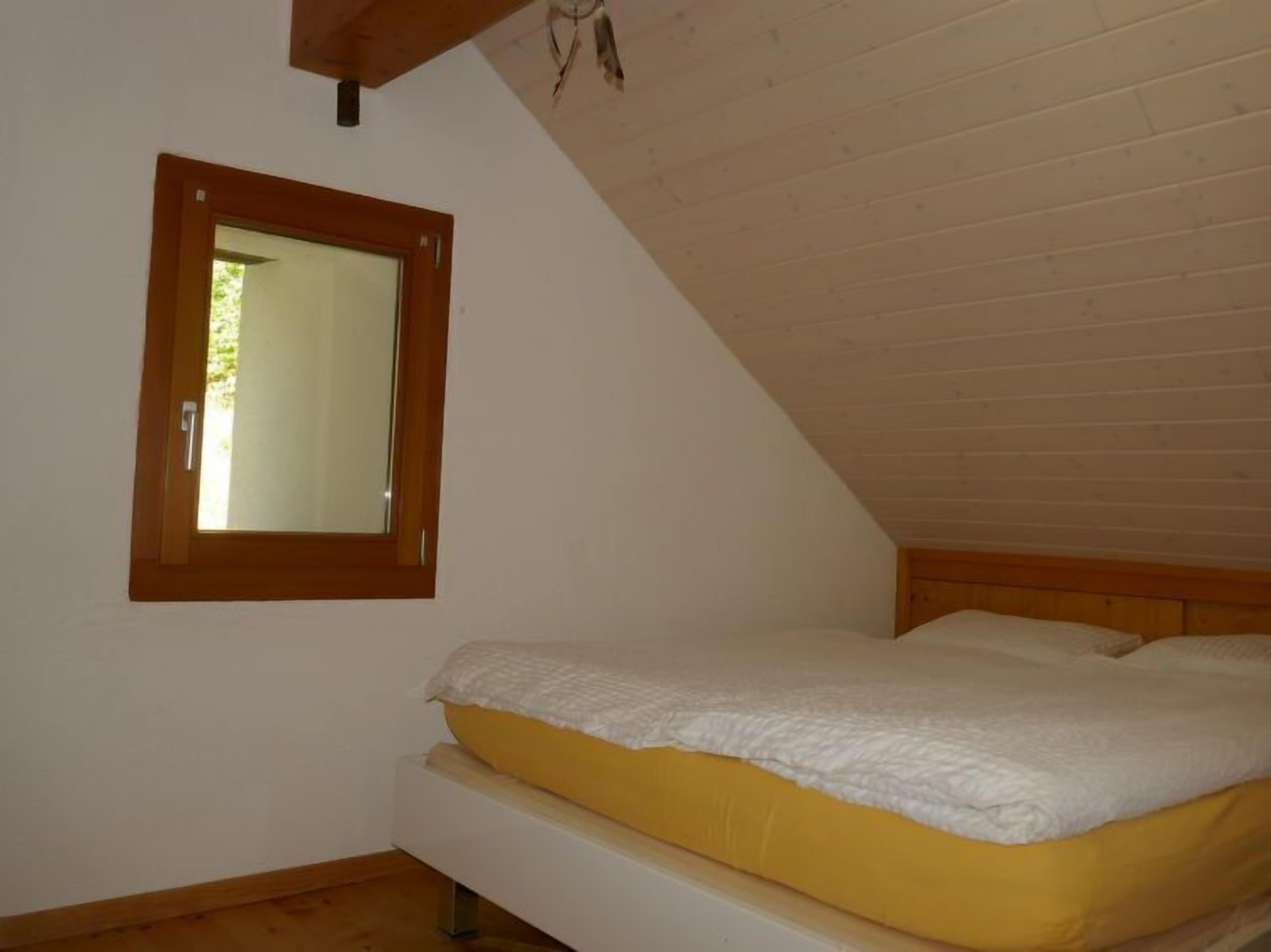 A bedroom with a sloped ceiling, a single bed with a yellow mattress and white bedding, and a small window with a wooden frame.