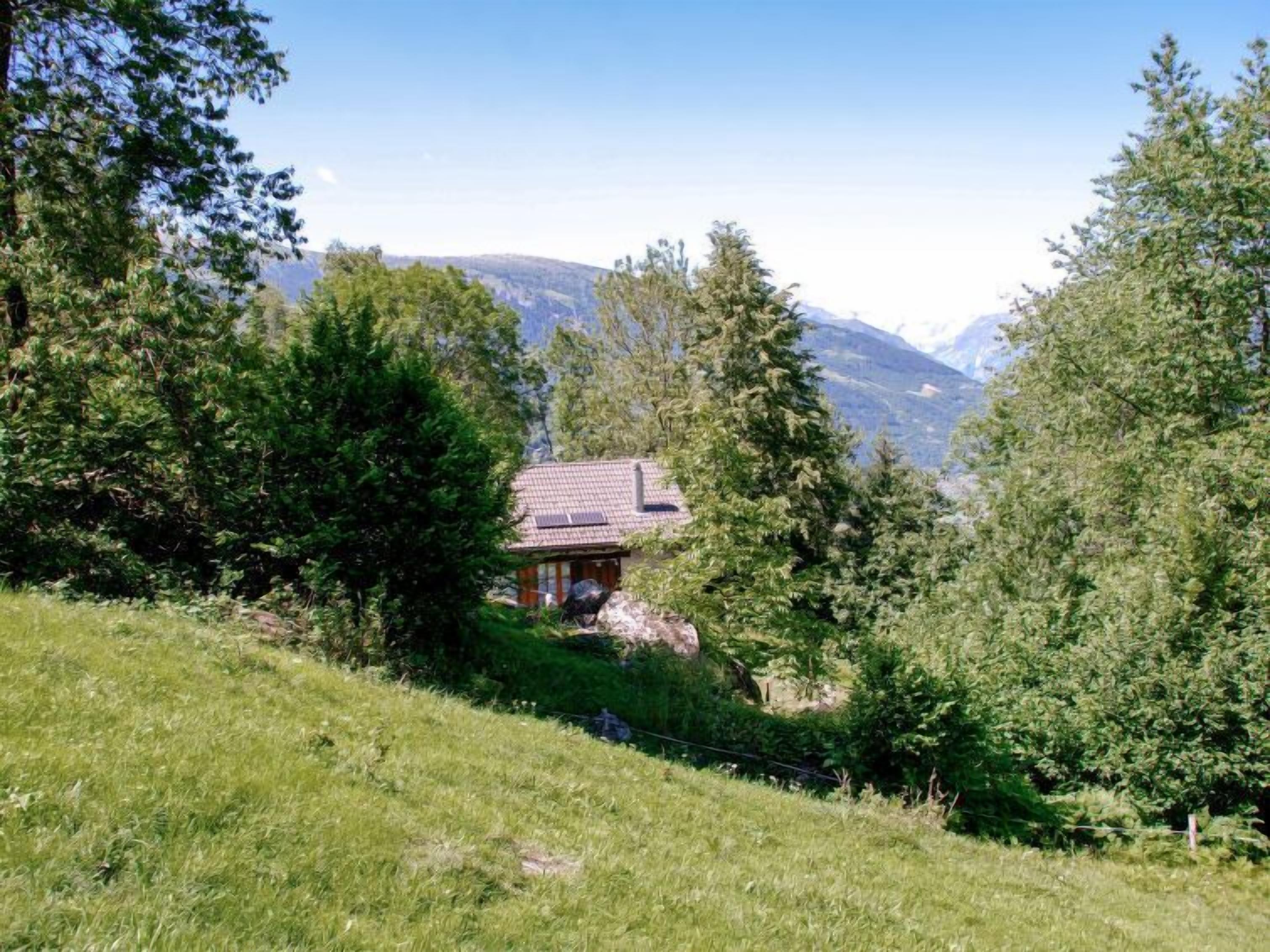 A small house with a tiled roof nestled among trees and grass, overlooking a valley with mountains in the distance.