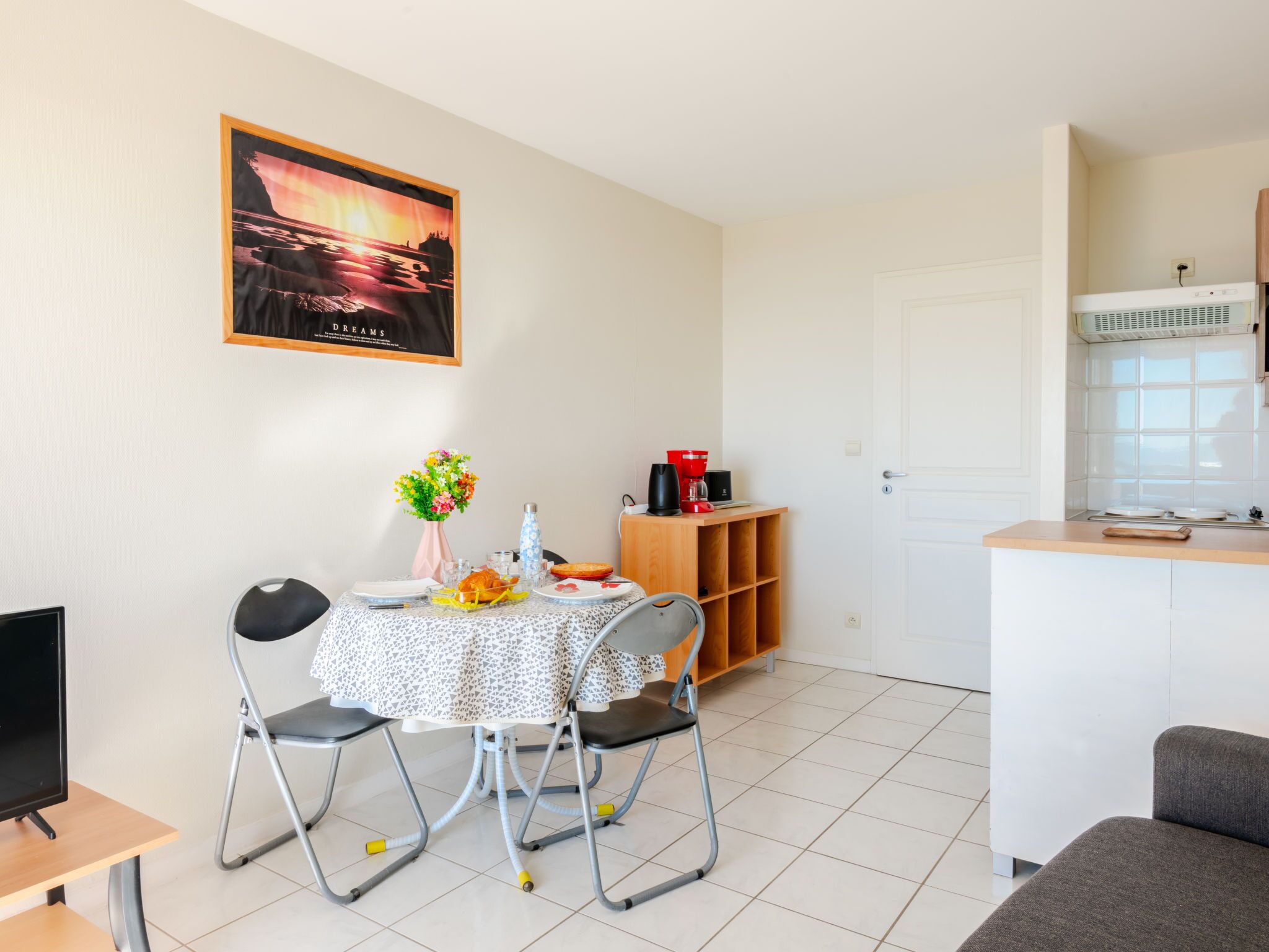 A compact kitchen with a dining area, a small table with chairs, a painting on the wall, and a window with a view.