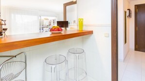 Eine moderne Küche mit einer Holzarbeitsplatte, einer Obstschüssel und einer gelben Vase. Zwei klare Glas-Barhocker und ein Metallstuhl sind ebenfalls vorhanden.