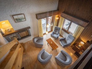 Um quarto com pé-direito alto, piso de madeira, lareira e um espaço de estar com quatro poltronas e uma mesa de centro.