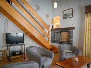 Uma sala de estar com escada de madeira, lareira, duas poltronas cinzas, mesa de centro de madeira e uma televisão sobre suporte.