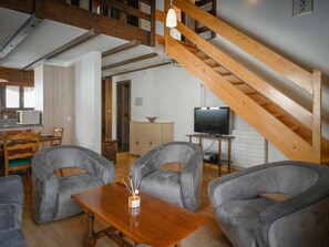 Una sala de estar moderna con sofás grises, una mesa de centro de madera y un televisor sobre un mueble de madera. Se ve una escalera con vigas de madera al fondo.