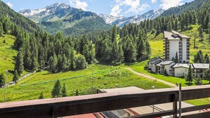 Una vista sulle montagne con un edificio a più piani e tetto piatto, immerso nel verde lussureggiante e con montagne innevate sullo sfondo.