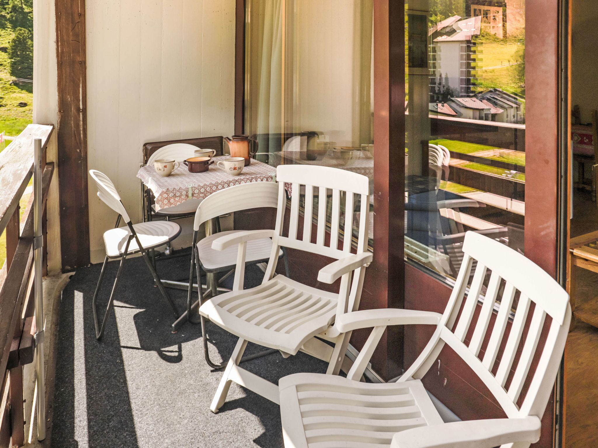 Ein Balkon mit weißen Plastikstühlen, einem kleinen Tisch mit Tischdecke und Blick auf eine grüne Landschaft durch eine Glastür.