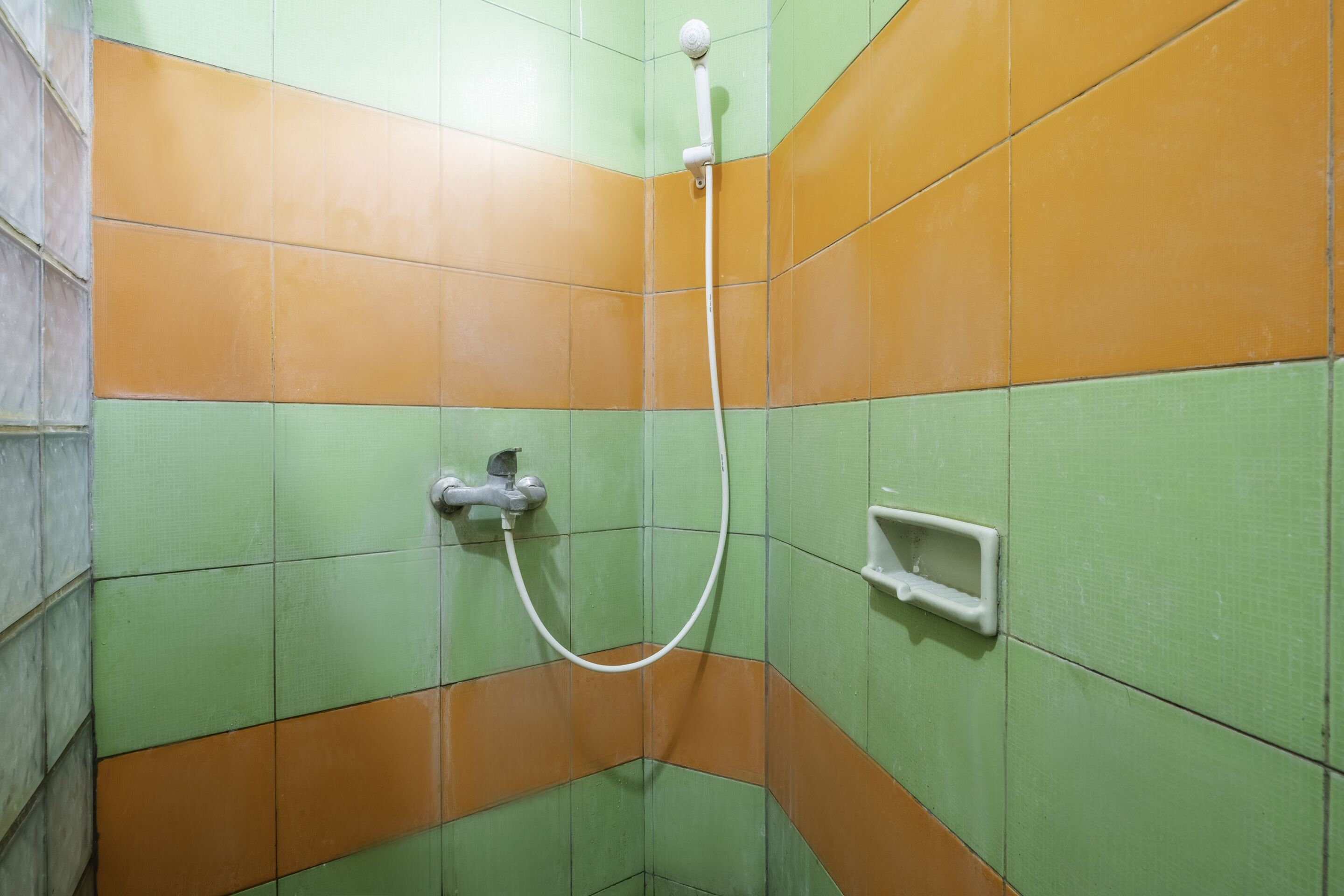 A corner of a bathroom with green and orange tiles, a white showerhead, and a small white soap dish.