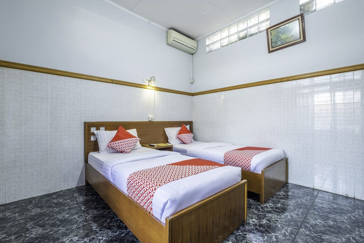Une chambre d’hôtel avec deux lits, une tête de lit en bois, une table de chevet, une lampe fixée au mur et une fenêtre avec des stores.