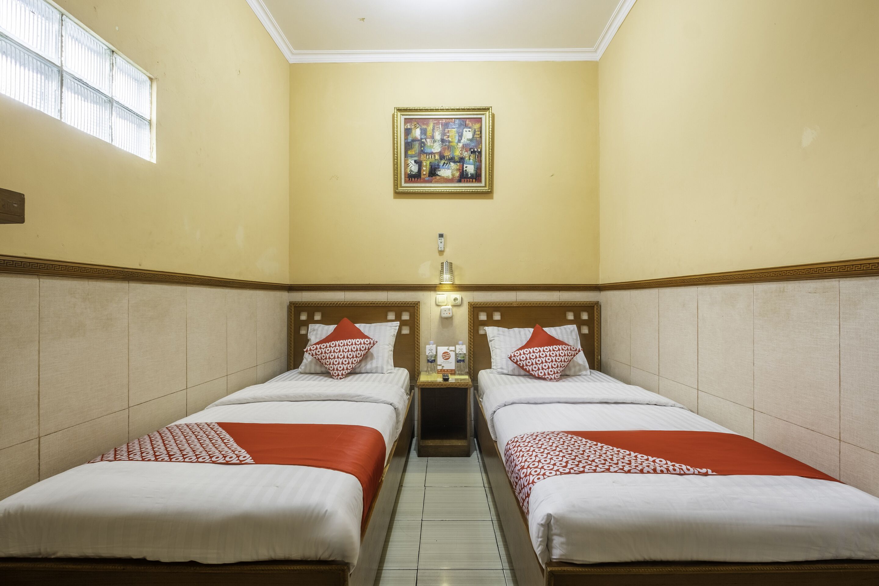 A hotel room with two beds, a nightstand, a wall-mounted light, and a window.