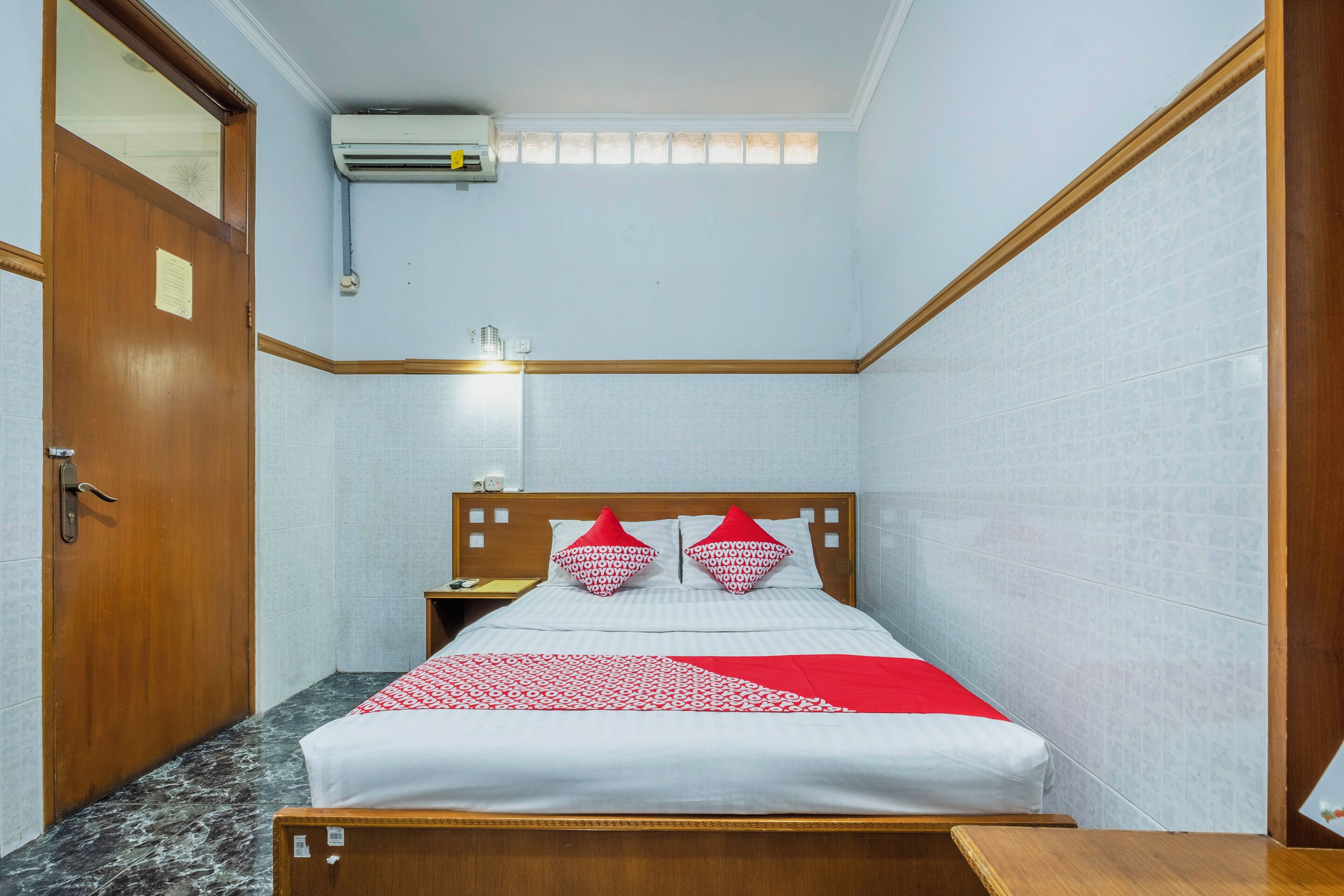 A hotel room with a bed, wooden headboard, a nightstand, a mirror, an air conditioning unit, and a door.