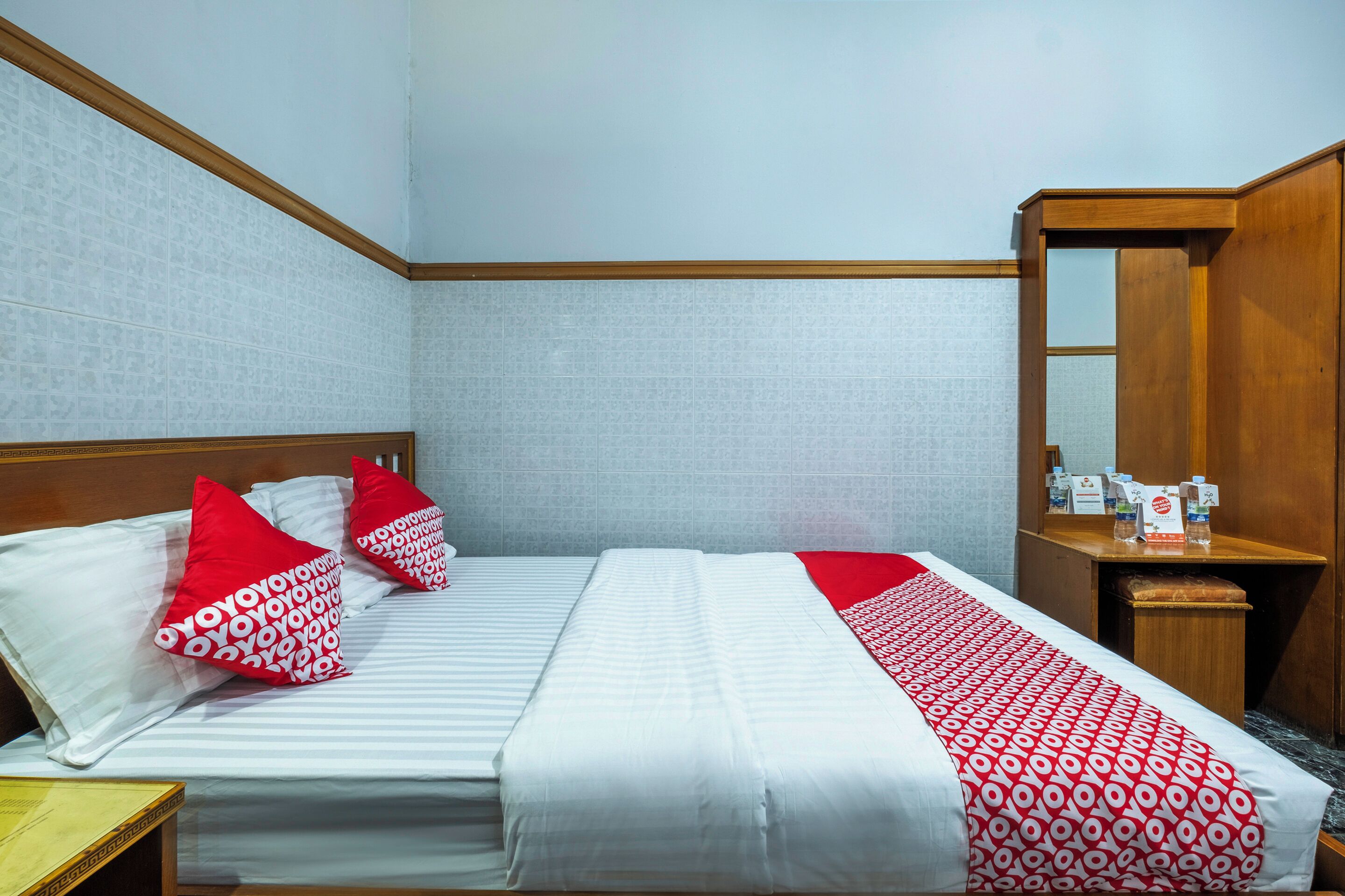 A hotel room with a bed, pillows, a bedside table, a mirror, and a wooden headboard.