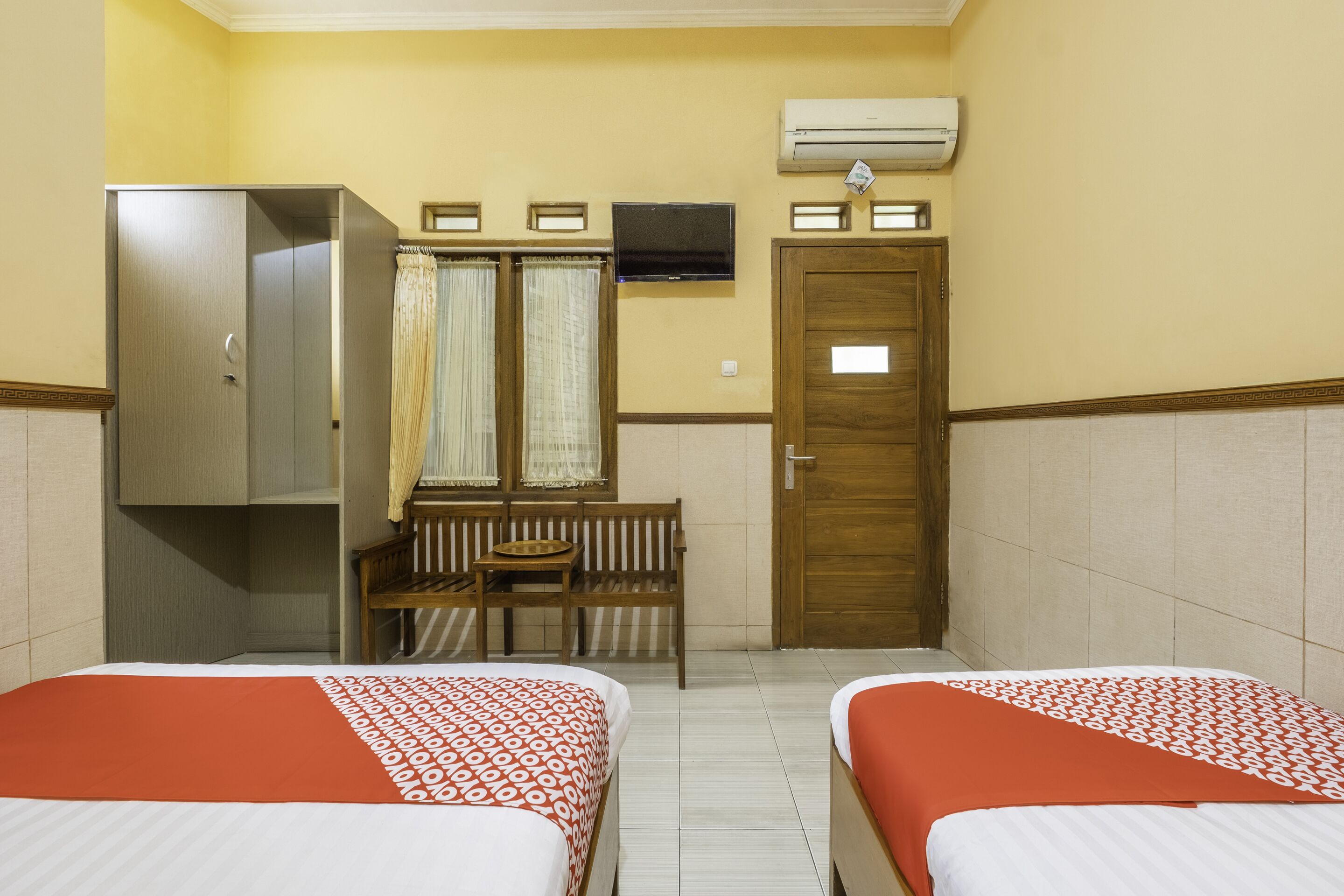 A hotel room with two beds, a wardrobe, a small table with a chair, a window with curtains, and an air conditioning unit.