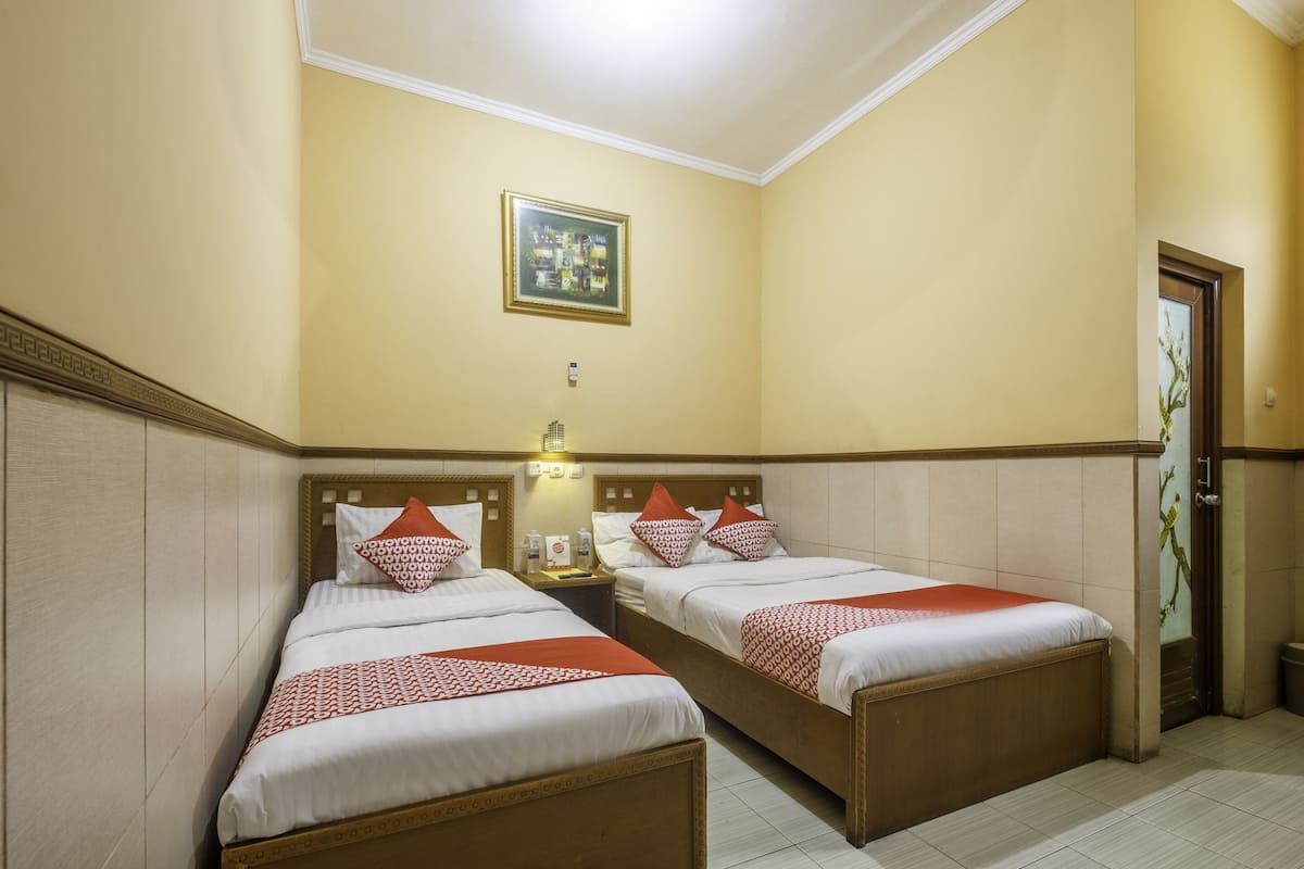 Une chambre d’hôtel avec deux lits simples, une table de chevet avec une lampe, un tableau encadré au mur et une porte ornée d’un motif décoratif.