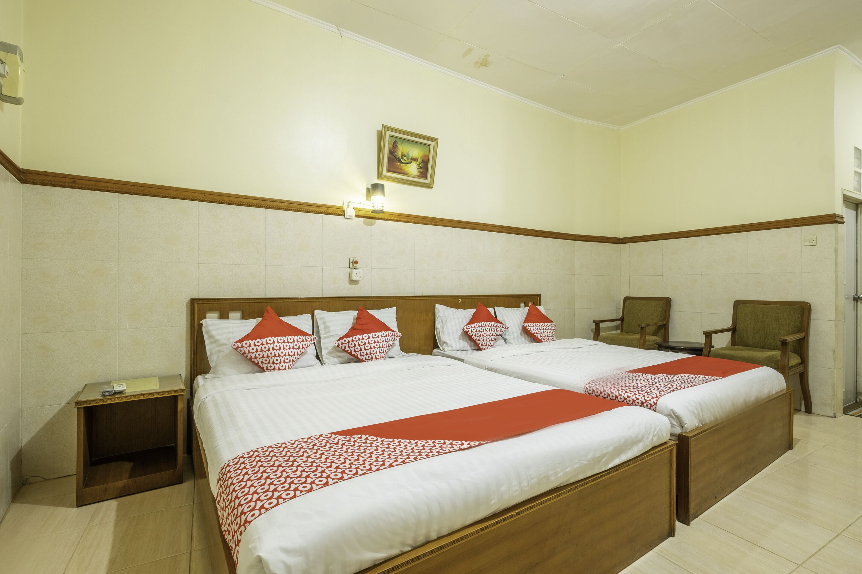 Camera d'albergo con due letti matrimoniali, un comodino, una sedia e un quadro appeso al muro.