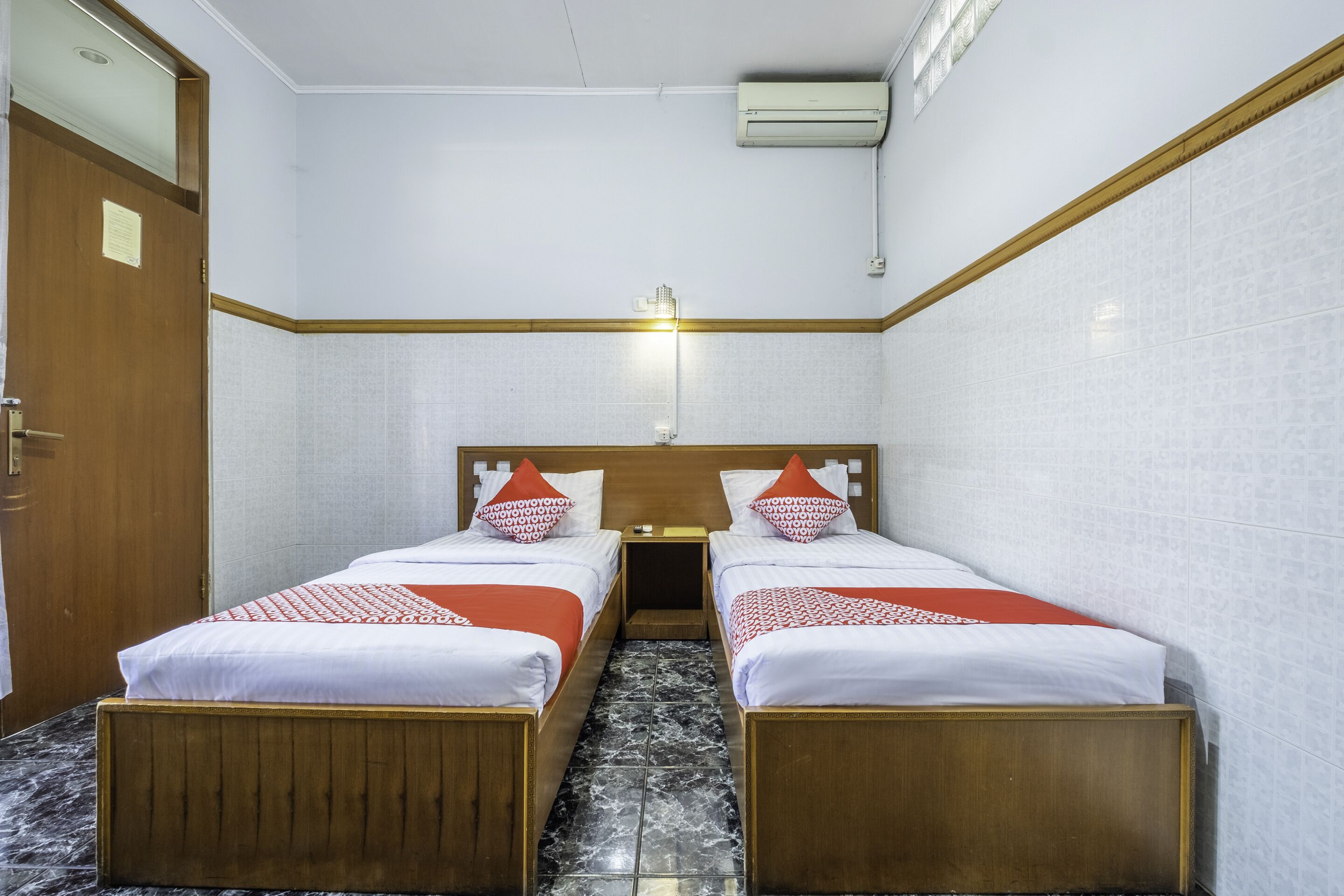 A hotel room with two beds, a wooden headboard, a nightstand, and a wall-mounted air conditioner.