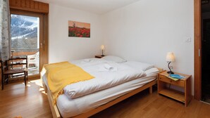 Een slaapkamer met een bed, houten nachtkastjes, een stoel, een raam met uitzicht op bergen en een schilderij aan de muur.