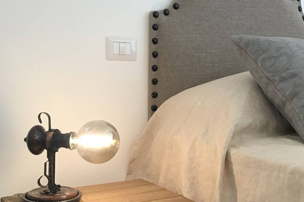 Een bed met hoofdbord, nachtkastje met lamp en stopcontact aan de muur.