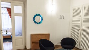 Um quarto com um armário de madeira, duas cadeiras pretas, um tapete azul, um espelho fixado na parede e um conjunto de portas venezianas.