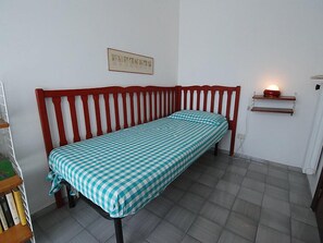 Uma cama de solteiro com colcha xadrez em quarto com piso de azulejos e cabeceira vermelha.