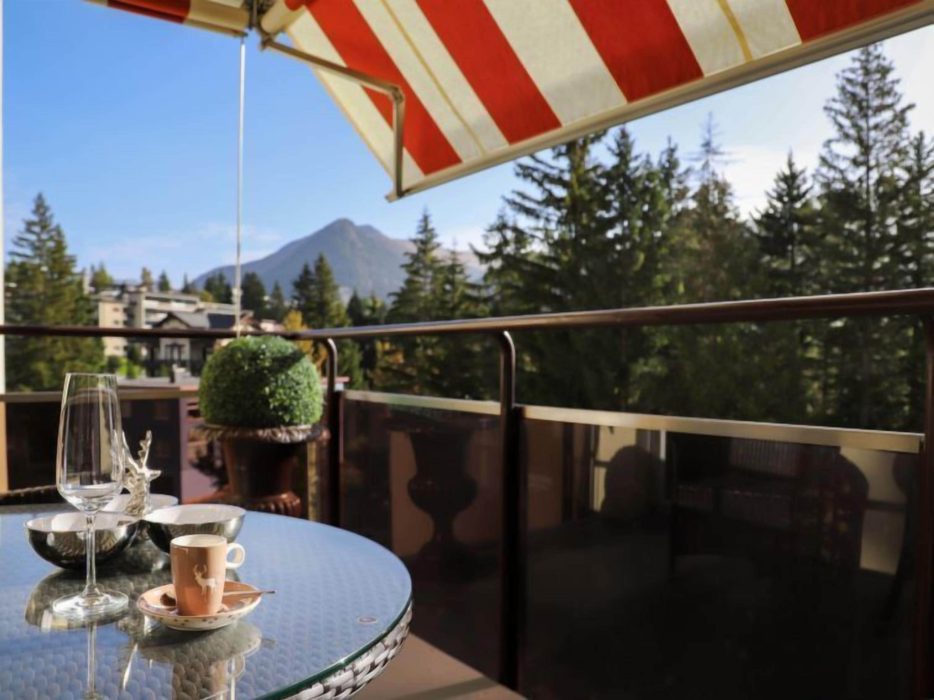 A round table with a glass top, a wine glass, and a cup on a balcony with a red and white striped awning.