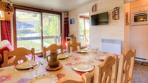 Un coin repas avec une table dressée pour quatre personnes, des chaises en bois, un téléviseur fixé au mur et une vue sur l’extérieur grâce à de grandes fenêtres.