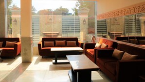 A modern lounge with red sofas, a central coffee table, and large windows offering a view of a garden and building facade. - Royal Residence Iffco Chowk (Gurugram)