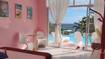 A bedroom with a bed, a small table, and chairs, a large window with a view of the ocean and a pool.