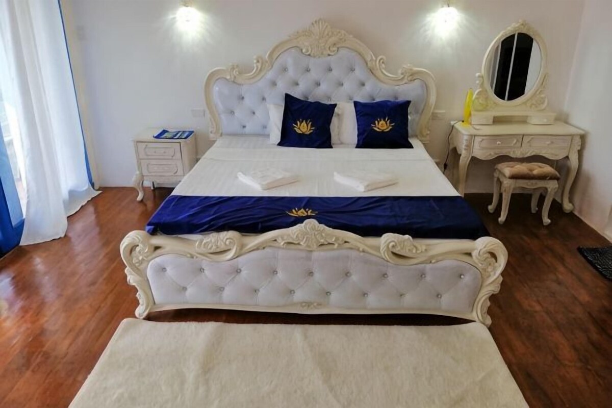Un dormitorio con una cama grande, una mesita de noche, un tocador con espejo y un taburete pequeño.