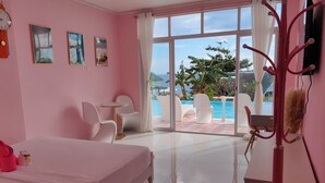 A pink-themed hotel room with a bed, two chairs, a small table, and a view of a pool and ocean.