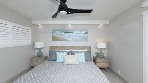 A bedroom with a bed, two nightstands, a ceiling fan, a wall-mounted painting, and a window with blinds.